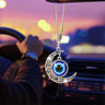 Silver tone crescent moon ornament with open scroll details and a hanging blue eye disc, shown below a car rearview mirror.