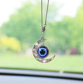 Silver tone crescent moon car ornament with open scroll details and a suspended blue eye disc, hanging in front of a windshield.