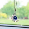 Silver tone crescent moon car ornament with open scroll details and a suspended blue eye disc, hanging in front of a windshield.