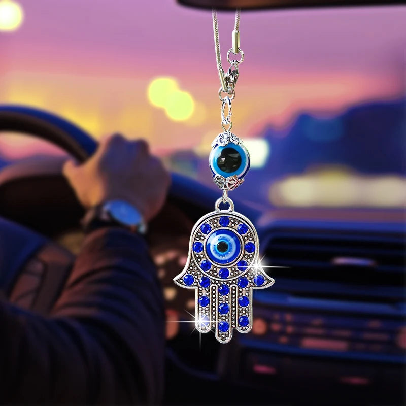 Hamsa hand evil eye ornament with blue accents, a silver tone frame and an upper eye bead, shown below a car rearview mirror.