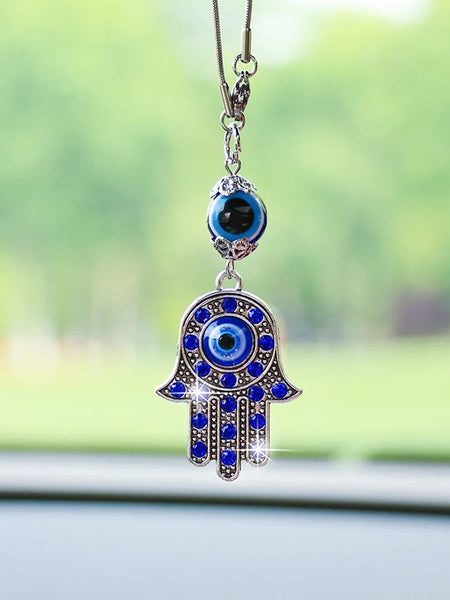Hamsa hand car ornament with blue accents, a silver tone frame and a separate eye bead above, hanging in front of a windshield.
