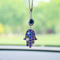 Hamsa hand car ornament with blue accents, a silver tone frame and a separate eye bead above, hanging in front of a windshield.