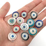 Twelve colorful oval Evil Eye connectors with clear accents and paired loops resting on a palm.