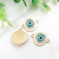 White oval Evil Eye connectors with gold tone frames and turquoise eye details, showing fronts and a grooved back.