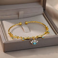 Gold tone cuff bracelet across a display box, with a blue eyelash eye charm and clear rectangular accents hanging over the edge.