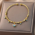 Gold tone cuff bracelet with a blue eyelash eye charm and clear rectangular accents, viewed from above on a beige cushion.