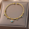 Gold tone cuff bracelet with a blue eyelash eye charm and clear rectangular accents, viewed from above on a beige cushion.