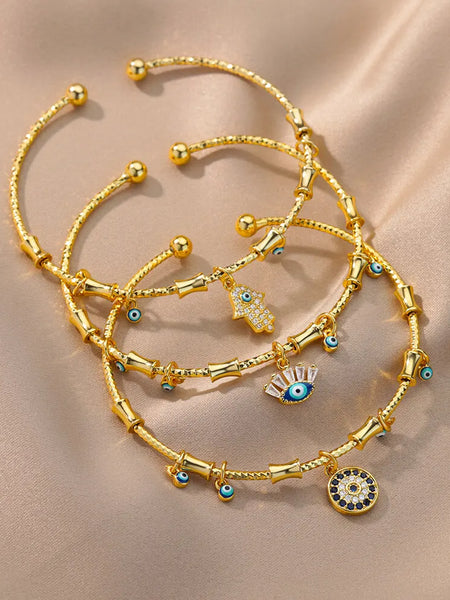 Three gold tone open cuff bracelet styles with Hamsa hand, eyelash eye and round charms overlapping on beige fabric.