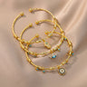 Three gold tone open cuff bracelet styles with Hamsa hand, eyelash eye and round charms overlapping on beige fabric.