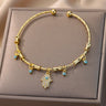 Gold tone Hamsa hand cuff bracelet viewed from above on a beige display cushion, with clear accents and small blue eye charms.