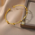 Gold tone round eye cuff bracelet angled beside a display box, showing the circular charm with dark blue and clear accents.