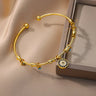 Gold tone round eye cuff bracelet angled beside a display box, showing the circular charm with dark blue and clear accents.
