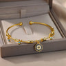 Gold tone cuff bracelet across a display box, with a round dark blue and clear-accented charm hanging over the front edge.