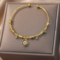Gold tone open cuff bracelet with a round blue and clear-accented charm, viewed from above on a beige cushion.