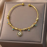 Gold tone open cuff bracelet with a round blue and clear-accented charm, viewed from above on a beige cushion.