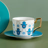 White cup with repeating blue Hamsa hands and circular eyes, an angular gold tone handle and a blue saucer