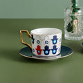 White cup patterned with navy, white and red Hamsa hands containing blue eyes, shown on a dark saucer