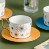 White cup with gold tone sunburst eye outlines and blue eye centers on a yellow saucer