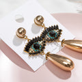 Gold tone teardrop earrings on a display card, with black lash accents and turquoise-colored eye details
