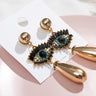 Gold tone teardrop earrings on a display card, with black lash accents and turquoise-colored eye details