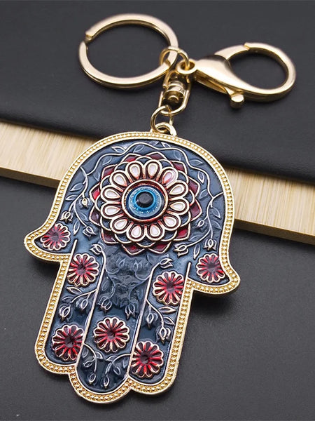 Blue Hamsa keychain with red flowers, a central blue Evil Eye and gold tone fittings, resting on a black book.