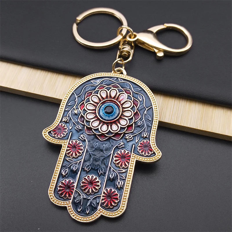 Blue Hamsa keychain with red flowers, a central blue Evil Eye and gold tone fittings, resting on a black book.