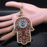 Brown floral Hamsa keychain with green accents and a blue Evil Eye, hanging against a sideways mannequin hand.