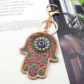 Brown and green floral Hamsa keychain with a blue Evil Eye and gold tone border, lying diagonally on printed pages.