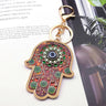 Brown and green floral Hamsa keychain with a blue Evil Eye and gold tone border, lying diagonally on printed pages.