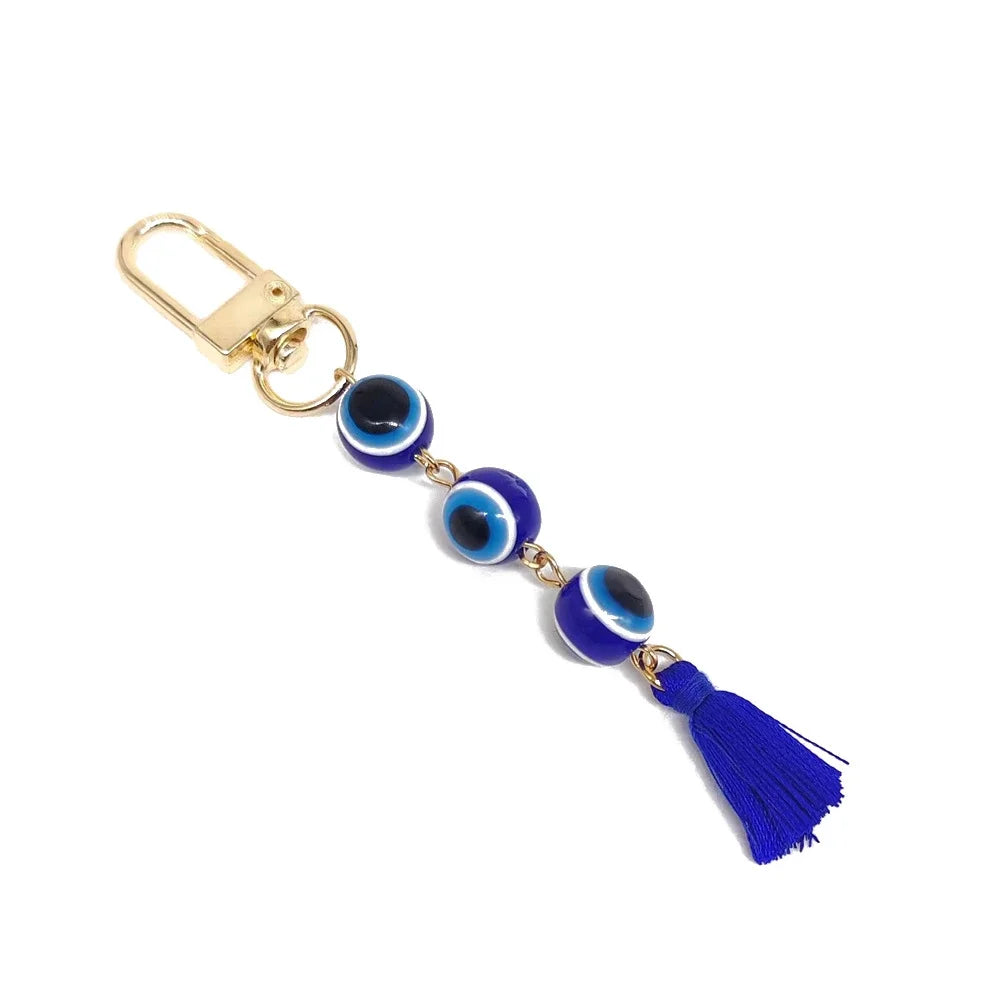 Dark blue evil eye keychain with three round beads, a blue tassel and a gold tone clip, arranged diagonally on white.