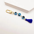 Light blue evil eye beads linked between a gold tone clip and a dark blue tassel, photographed diagonally beside a white block.