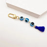Light blue evil eye beads linked between a gold tone clip and a dark blue tassel, photographed diagonally beside a white block.