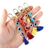 Four evil eye tassel keychains resting across a hand, with dark blue, red, multicolored and light blue beads and gold tone clips.