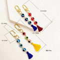 Four evil eye tassel keychains with measurement lines and supplier annotations showing approximately 10.5 cm, 11.5 cm, 3.5 cm, 1.3 cm and 6 g.