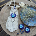 Off-white macrame wall hanging with a blue eye pendant, triangular knot panels and long pointed fringe on a round woven backdrop.