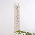 Off-white macrame hanging with seven blue eyes and a leafy branch overlapping the lower fringe