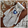 Angled view of the side-tassel evil eye hanging showing off-white knots around a blue pendant and separate twisted side cords.