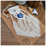 Off-white evil eye macrame hanging with a round blue pendant, two side tassels and pointed central fringe, laid diagonally on a brown board.