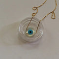 Round white evil eye pendant with a turquoise blue ring and black center on a gold tone chain.