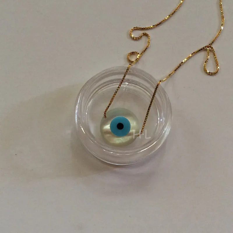 Round white evil eye pendant with a turquoise blue ring and black center on a gold tone chain.