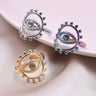 Gold tone, silver tone and rainbow evil eye rings displayed around a pale dish, showing the open oval faces and rounded border accents.