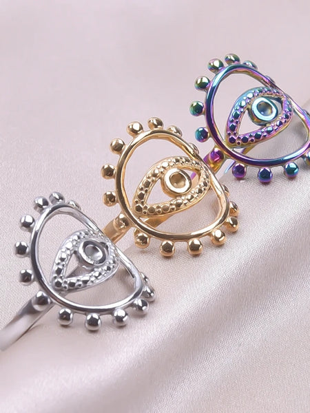 Silver tone, gold tone and rainbow evil eye rings arranged diagonally on pale fabric, each with a beaded oval frame.