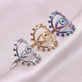 Silver tone, gold tone and rainbow evil eye rings arranged diagonally on pale fabric, each with a beaded oval frame.