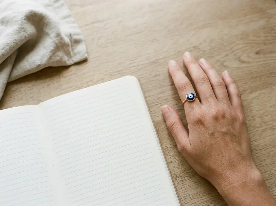 Relaxed hand with a single evil eye ring beside a blank journal and folded fabric on a beige tabletop.