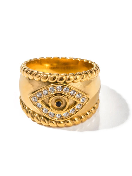 Gold tone wide band evil eye ring with clear accents, a black center and rope-style edges on a white background