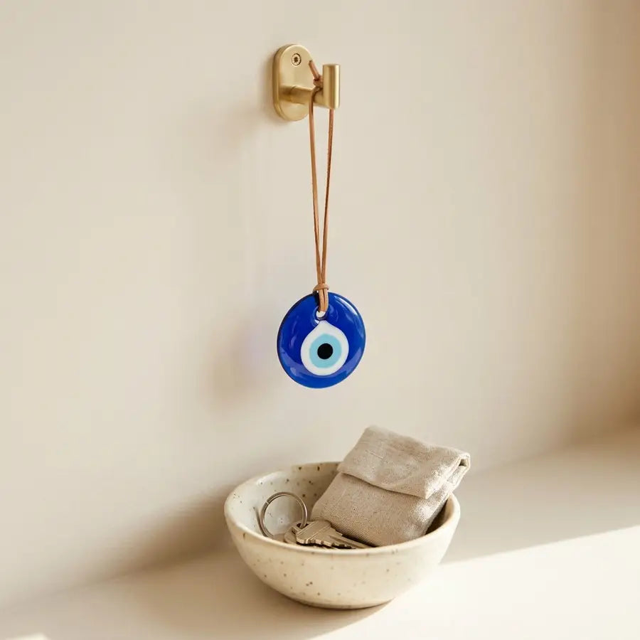 A single evil eye charm hangs above a ceramic bowl with keys and a linen pouch, with the blue-and-white eye sharply visible.
