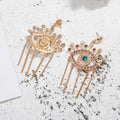 Gold tone evil eye fringe earrings showing an embellished front, textured reverse and straight posts on a speckled surface.