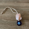 Evil eye wall hanging with a blue teardrop, pink stone accent and cream cord loop laid on a wood surface