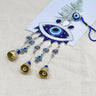 Blue evil eye wall hanging with Hamsa charms and three gold tone bells on pale fabric beside a fern print