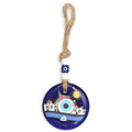 Blue evil eye wall hanging with white village buildings, flowers, a sailboat and a yellow sun